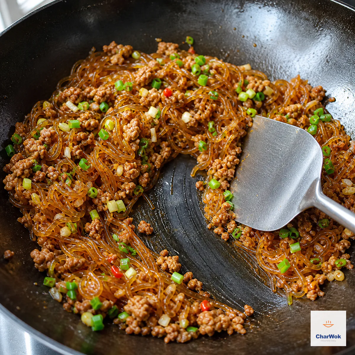 A wok spatula leaves a clean, clear dark-metal streak through glossy red glass noodles and small pork crumbles with no standing broth