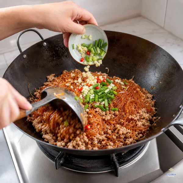 Fresh minced garlic, scallion greens, and a few red chili rings fall into glossy red glass noodles and distinct pork crumbles in an off-heat wok