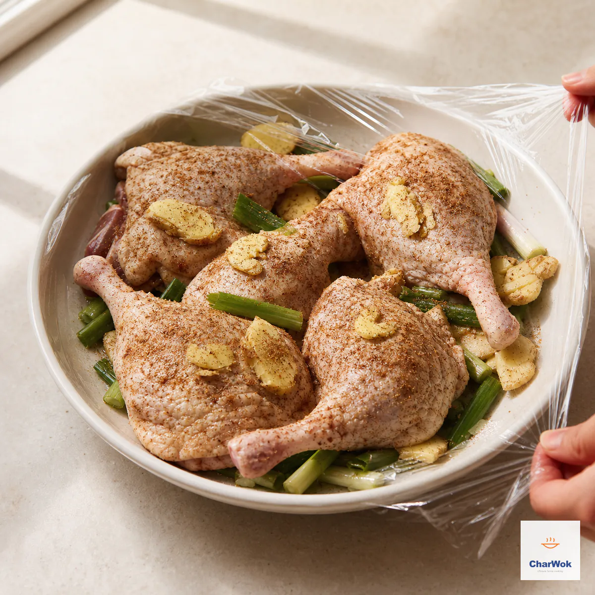 Four dry-spiced raw duck legs rest on a bed of scallion and ginger in a deep plate under clear wrap for the overnight cure
