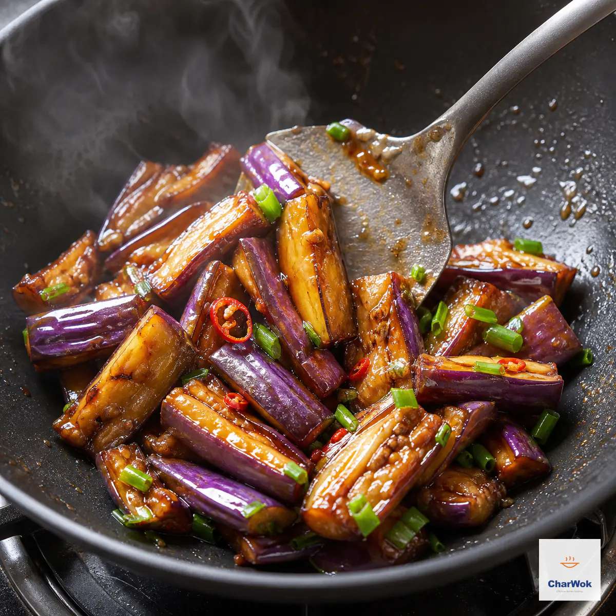 A spatula folds soft intact eggplant through a thin glossy plum glaze with bright scallion greens and red chili rings, leaving almost no liquid in the wok