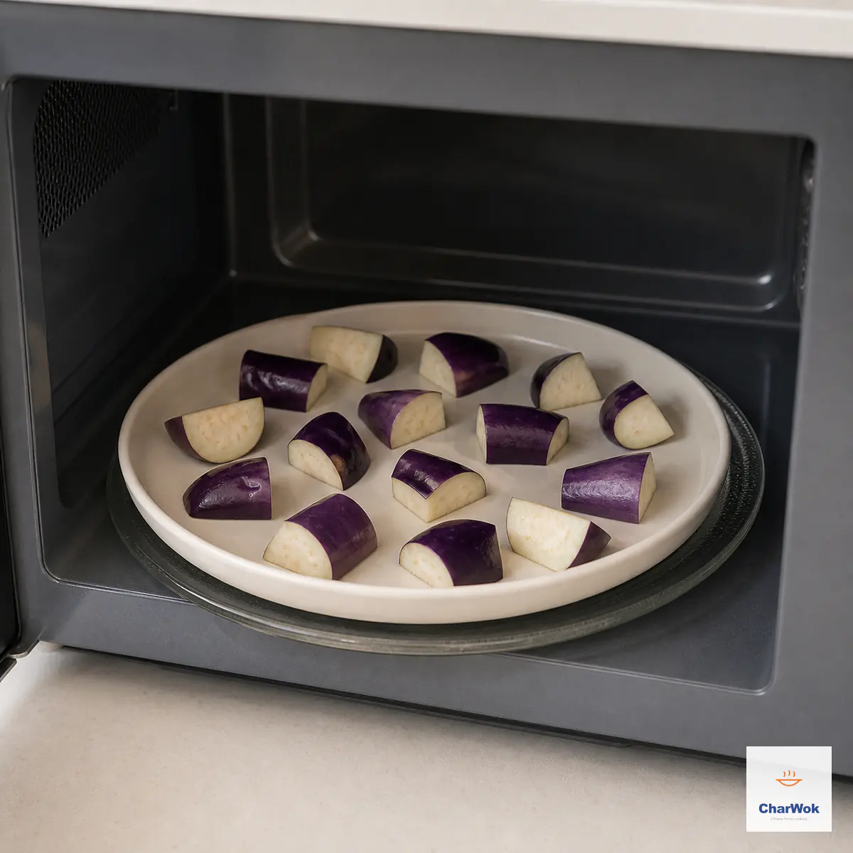 Skin-on eggplant chunks sit in one uncrowded layer with visible gaps on a ceramic plate inside an open microwave