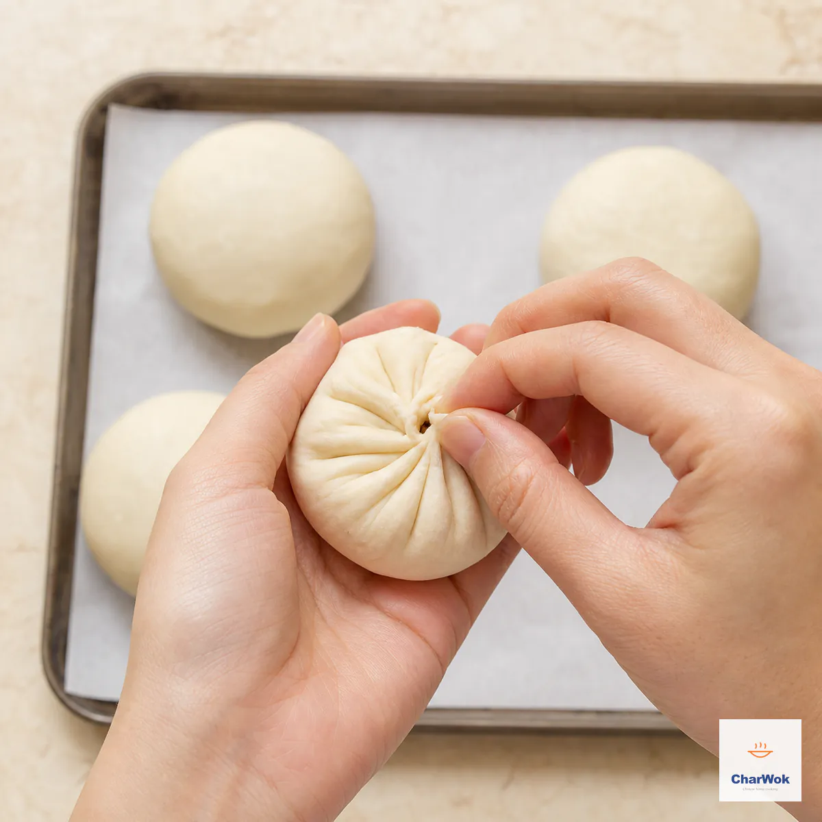 Two hands pinch the final tiny opening in a finely pleated round lamb bao while smooth sealed buns rest seam-side down on parchment behind it