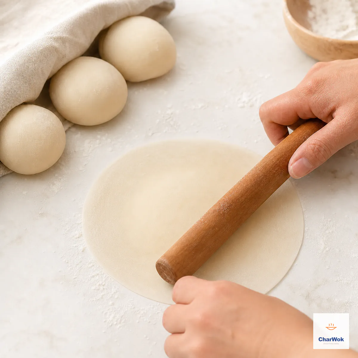 A short wooden rolling pin thins the edge of one seamless round yeast-dough wrapper while its center remains slightly thicker