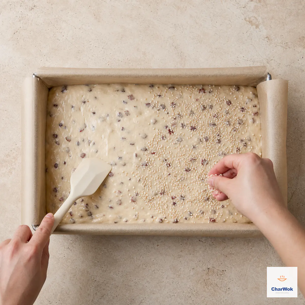 Two hands level date-studded batter and scatter white sesame in a wide rectangular pan lined across the bottom and all four sides with parchment