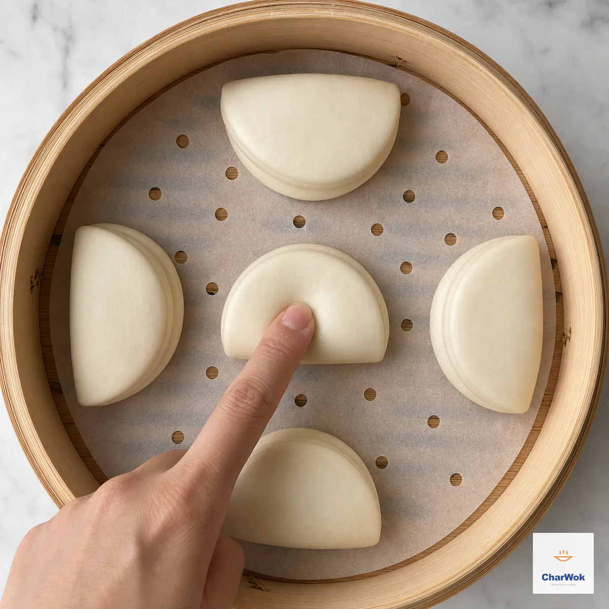 Five plump raw folded bao buns proof with space between them in a lined bamboo basket while a fingertip tests the centre bun