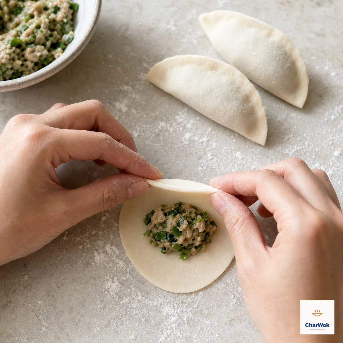 Dry fingers press the clean un-oiled rim of a modestly filled round wheat wrapper into a flat half-moon seam while two sealed dumplings sit nearby
