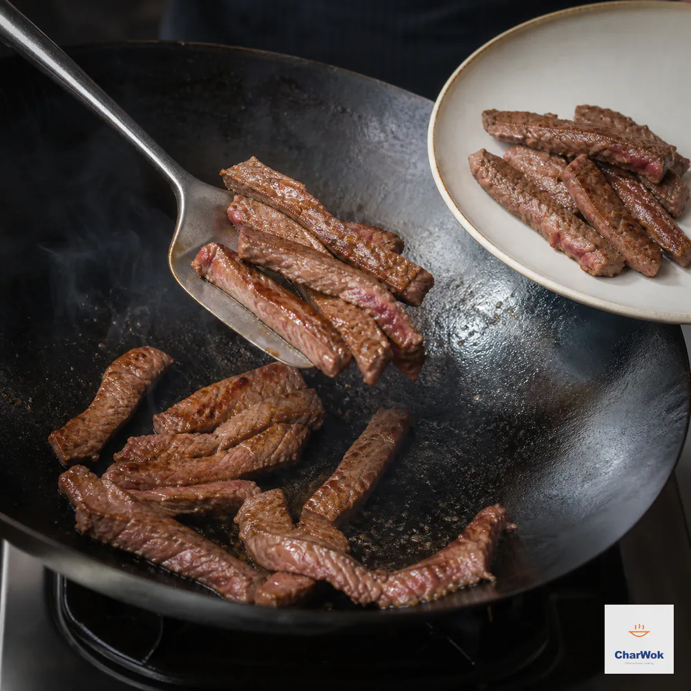 A metal wok spatula lifts browned beef strips from a very hot uncrowded wok while the cut edges still show a faint pink center