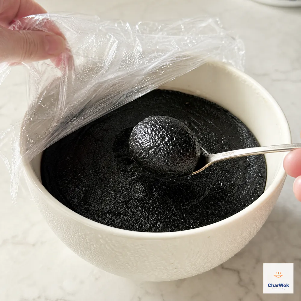 Black sesame paste chills under plastic wrap pressed directly onto its surface while a rounded scoop beside it holds a clean shape