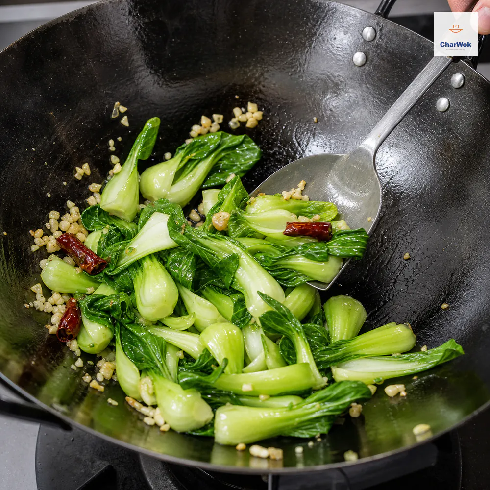 A broad wok spatula rapidly scoops bright drained bok choy with pale-gold coarse garlic and three dried chilies across an exposed wok bottom with no water pool