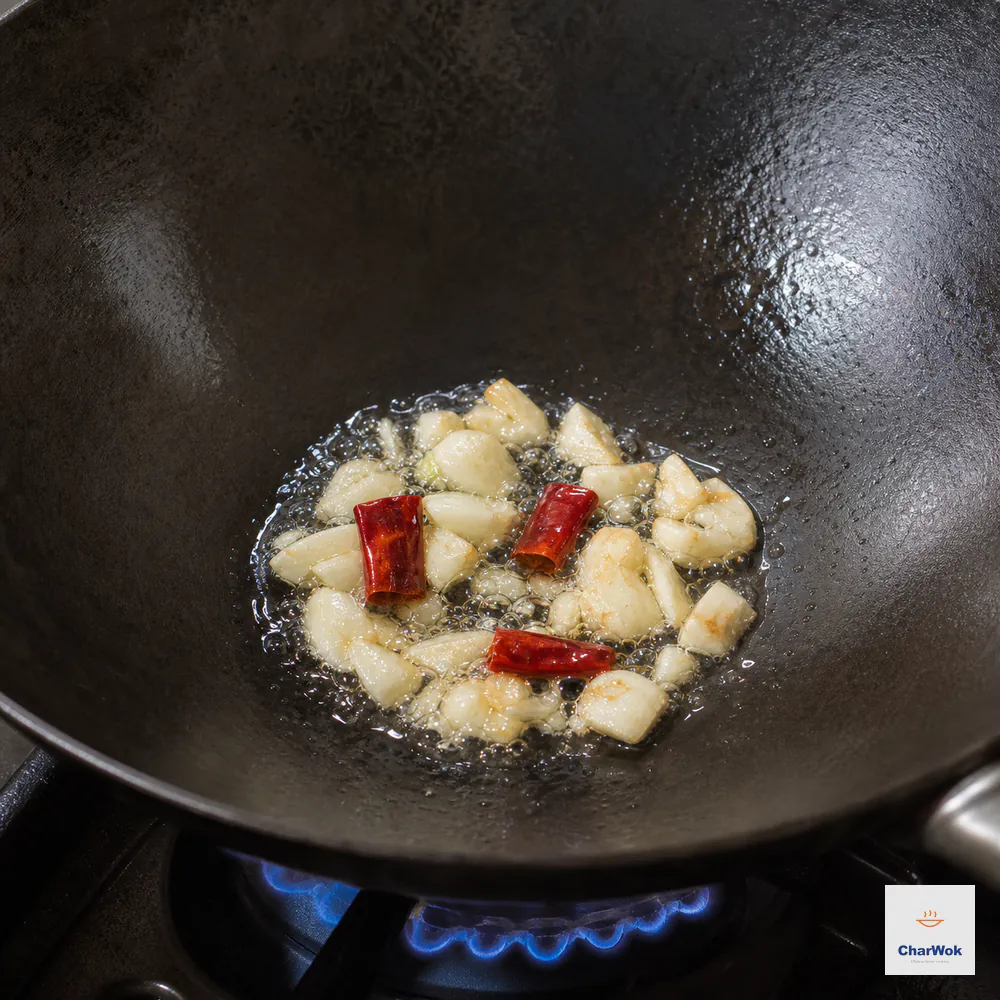 Coarsely smashed garlic and three dried red chili segments sizzle in a small pool of neutral oil over a blue flame while the garlic edges remain only faintly gold