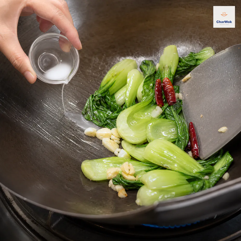 A tiny cup pours a hair-thin cornstarch slurry stream onto bare hot wok metal beside bright bok choy as a spatula forms a clear clinging gloss without a sauce pool