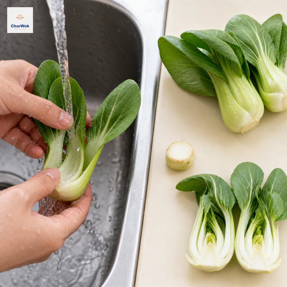 Hands rinse the grit-catching inner cup of raw baby bok choy beside a trimmed hard root base, whole small heads, and one larger head halved lengthwise