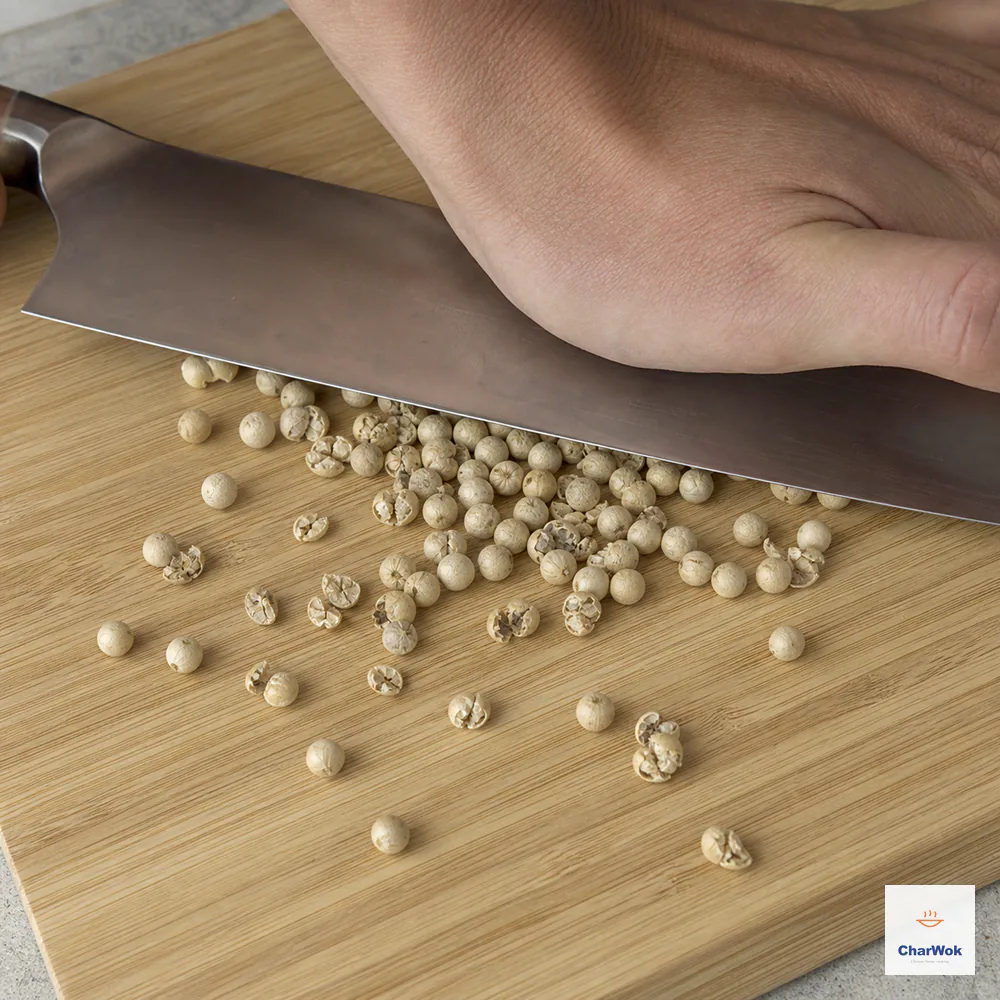 The flat side of a heavy knife cracks pale whole white peppercorns into coarse split pieces while the heel of one hand presses safely on the blade