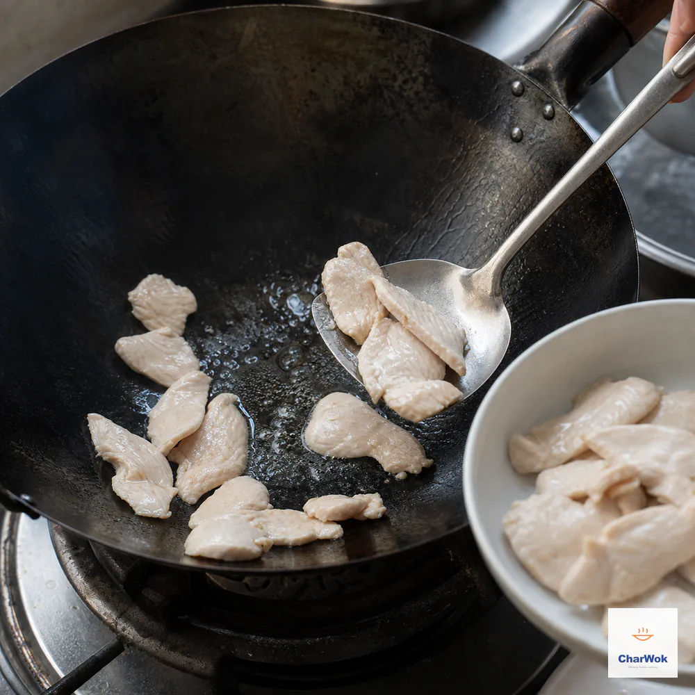 A wok spatula transfers partially seared chicken breast slices from a lightly oiled carbon-steel wok to a clean plate before the centers are cooked