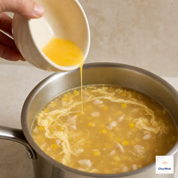 A very thin thread of beaten egg falls into barely simmering chicken corn soup as long pale egg ribbons set across the calm surface