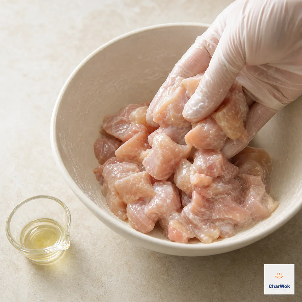 A gloved hand lifts raw chicken pieces from a cream bowl after cornstarch, salt, and water form a sticky coating with no free liquid at the bottom