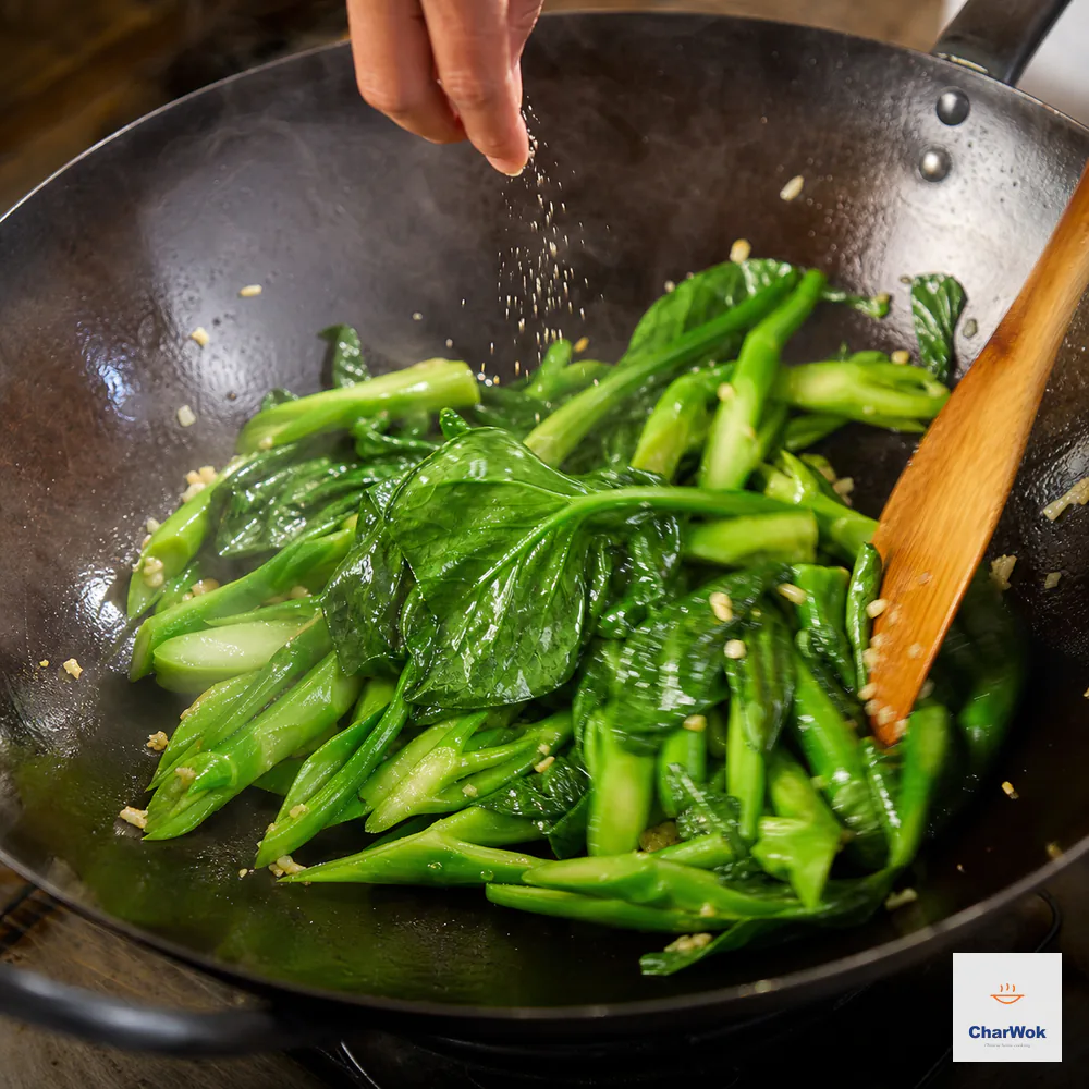 A small pinch of sugar and salt falls over broad gai-lan leaves and diagonal stems during the final fast toss in a dry wok