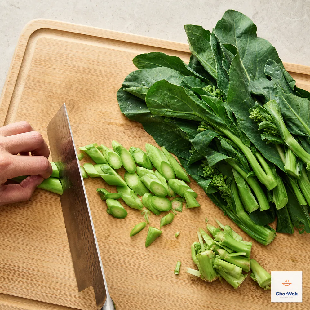A cleaver cuts raw gai-lan stems diagonally into thin pieces while broad leaves and discarded hard ends remain in separate piles
