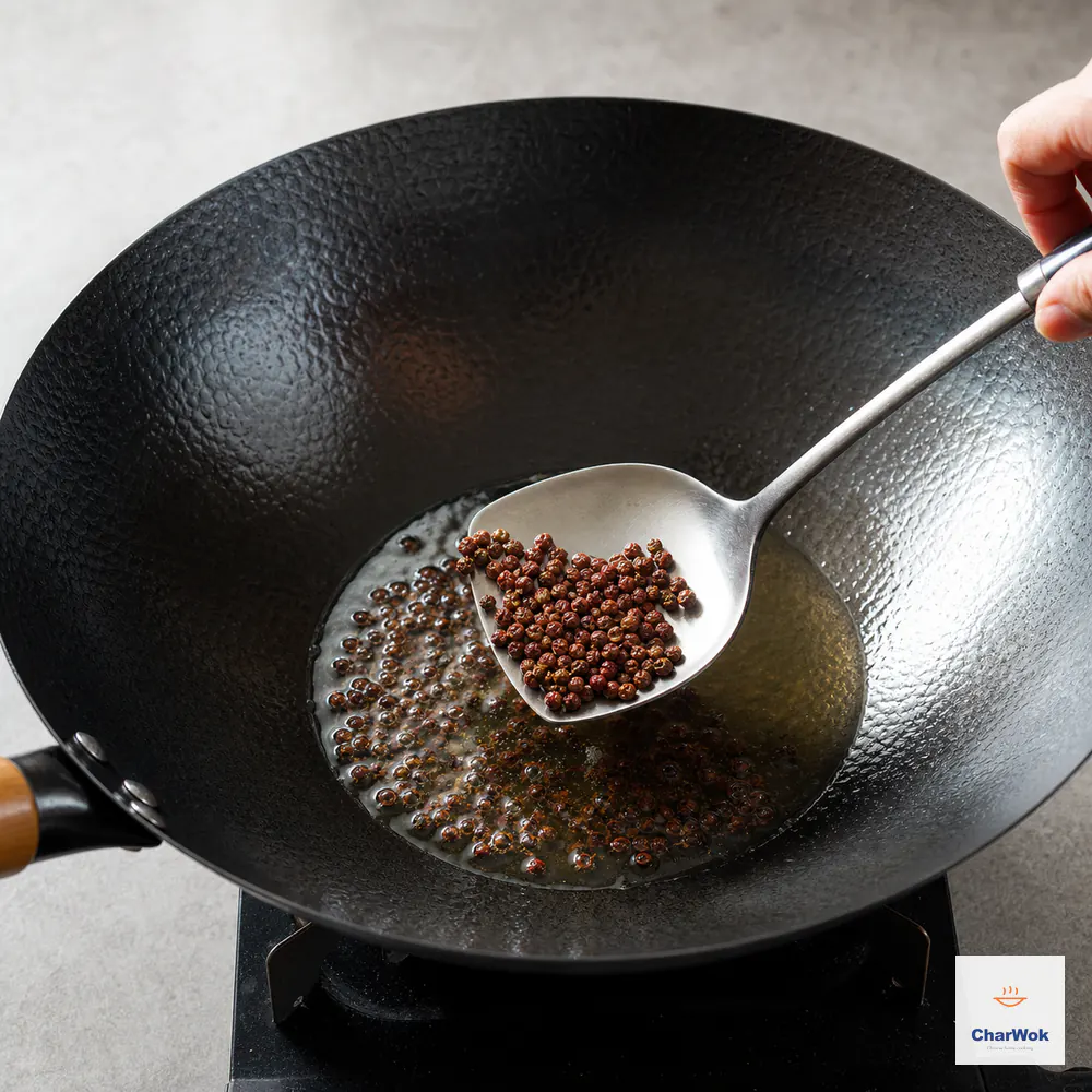 A metal wok spatula lifts reddish-brown Sichuan peppercorns from shimmering oil as tiny bubbles form in a hammered carbon-steel wok