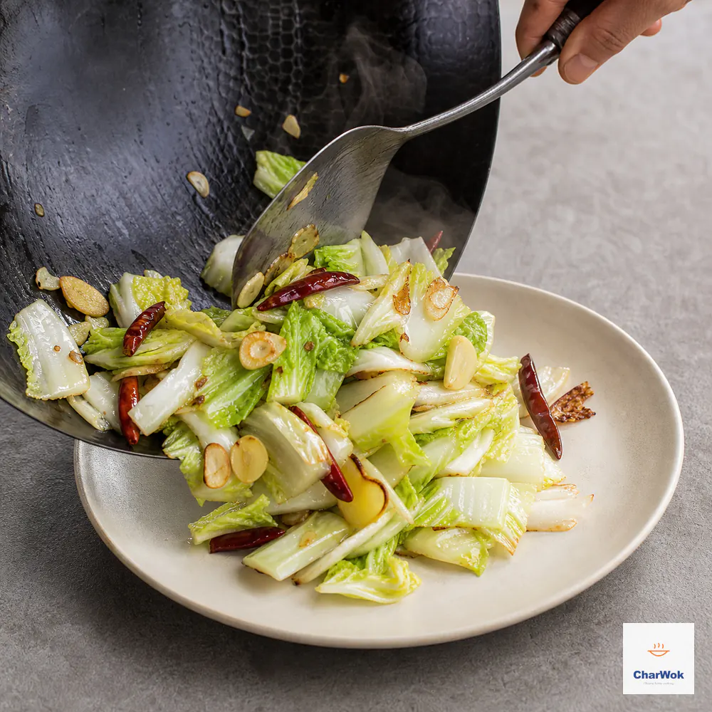 A wok spatula slides glossy but dry cabbage with garlic, ginger, and dried chillies onto an ivory plate with no liquid pool