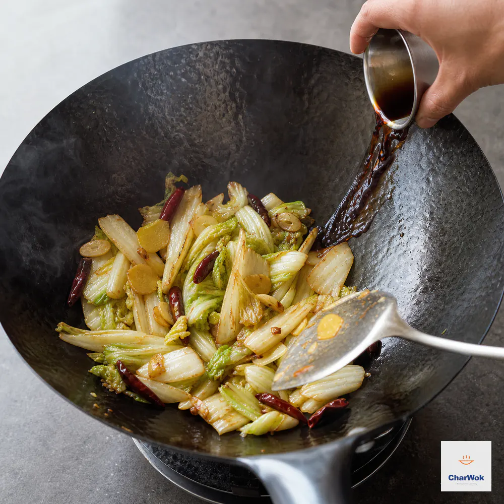 A small cup pours a restrained soy-and-vinegar seasoning down the bare hot wok rim beside wilted cabbage with lightly browned edges