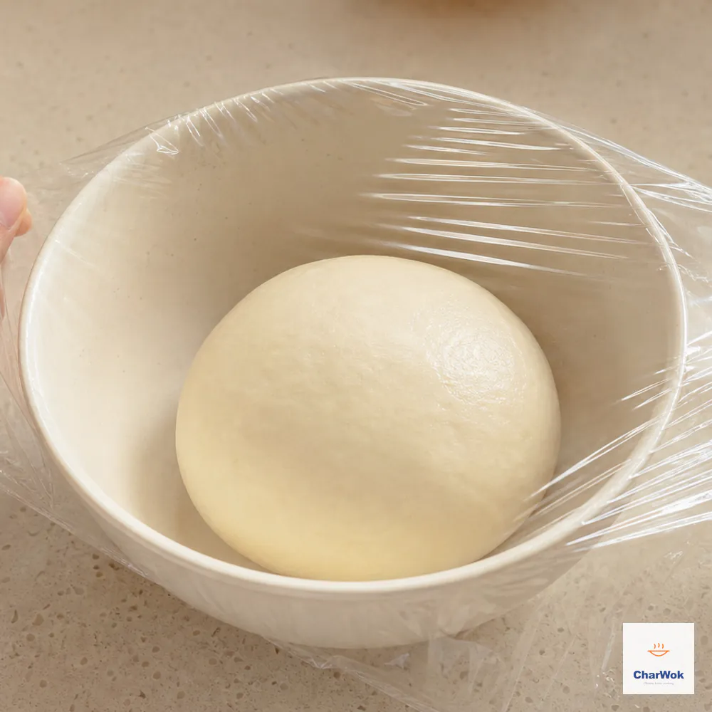 A soft smooth ball of warm hot-water dough is being covered tightly with clear film in a cream ceramic bowl