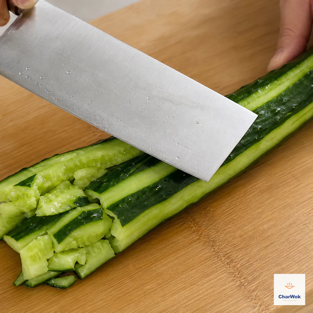 A large cucumber with alternating peeled stripes is being firmly smashed with the flat side of a Chinese cleaver, forming rough split pieces rather than neat slices