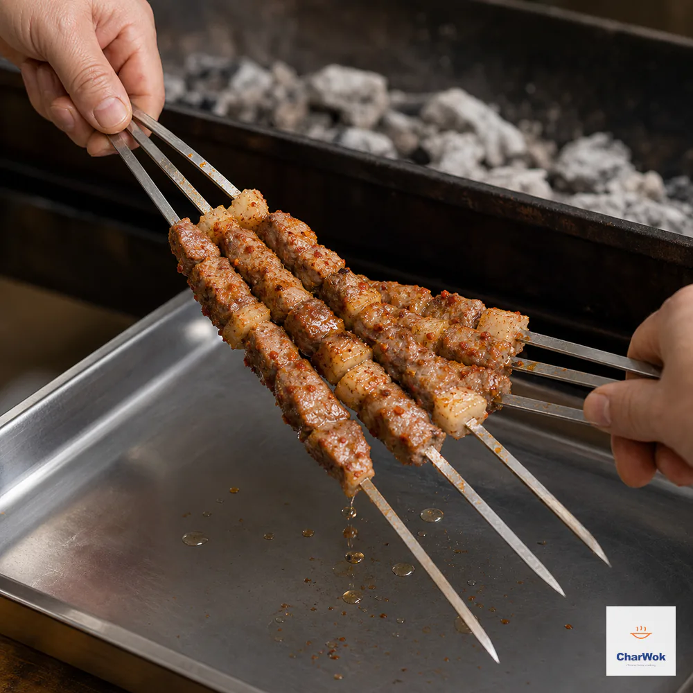 Two hands press and rub one bundle of three browned lamb skewers against another bundle of three above a metal catch tray as rendered fat drips