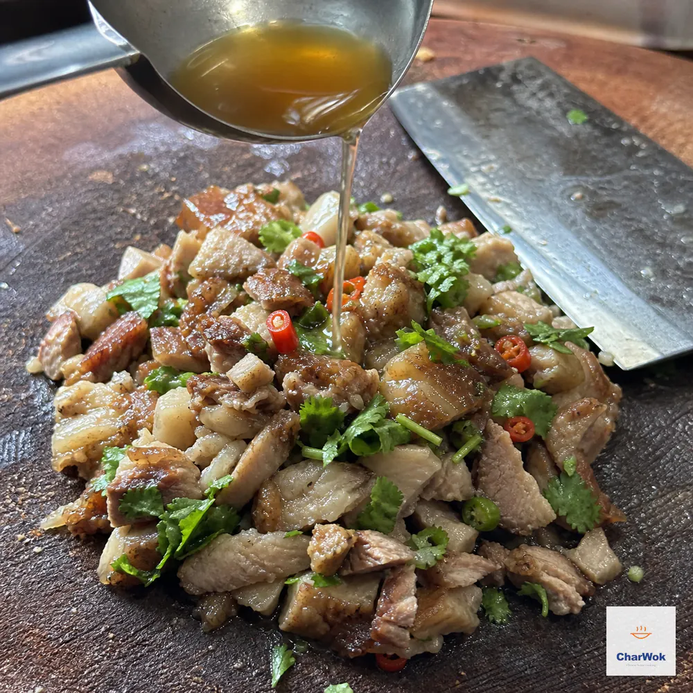A small ladle pours warm amber braising broth over irregular chopped pork belly, cilantro, and fresh chili beside a resting cleaver, with no liquid pool