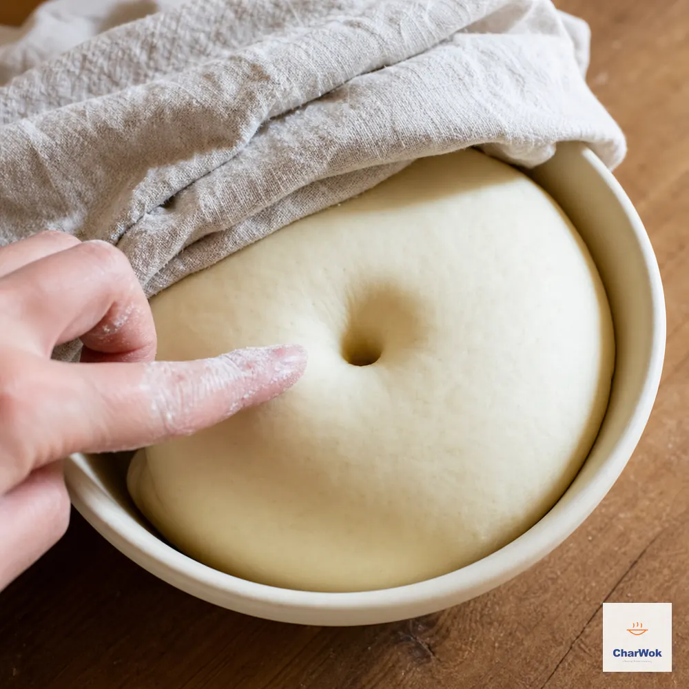A flour-dusted finger has left one deep round hole in modestly doubled dense dough, and the hole stays open without the dough collapsing