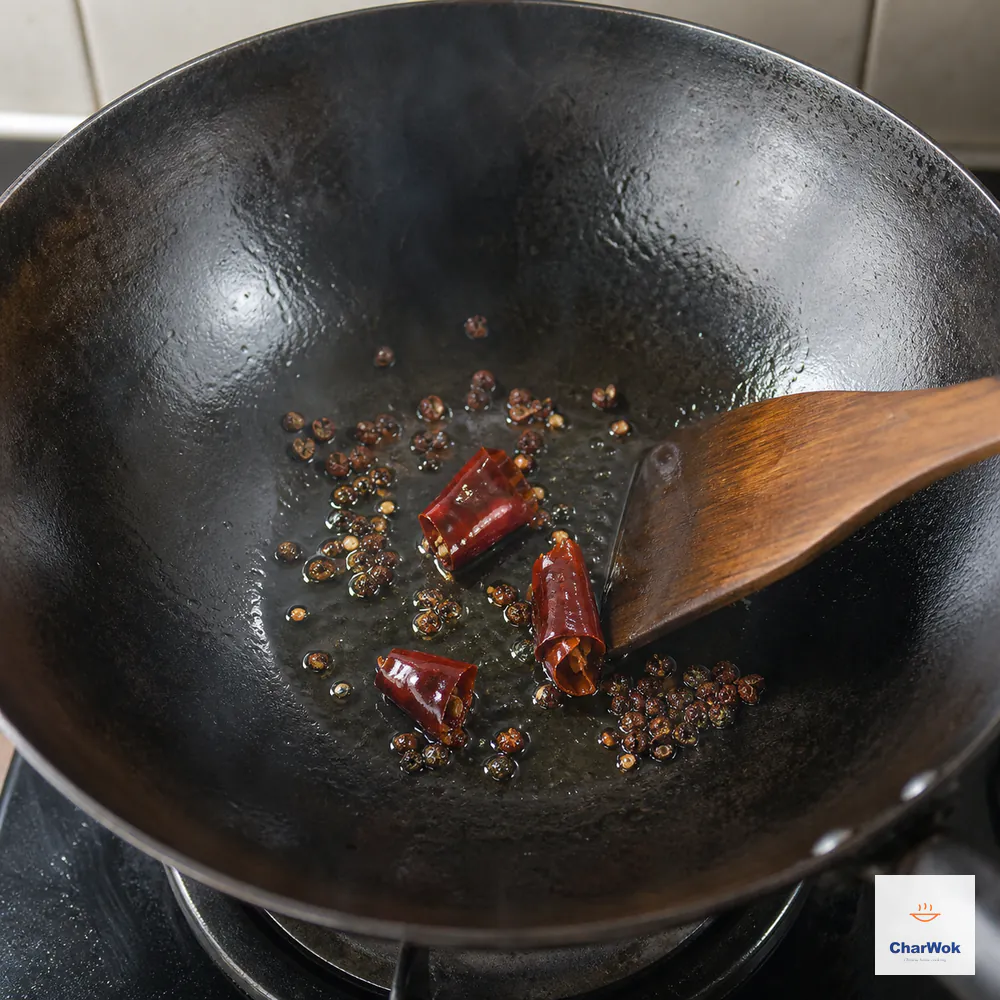Three dried chili sections and whole red Sichuan peppercorns sizzle in a thin oil sheen as a wooden wok spatula keeps them moving before they blacken