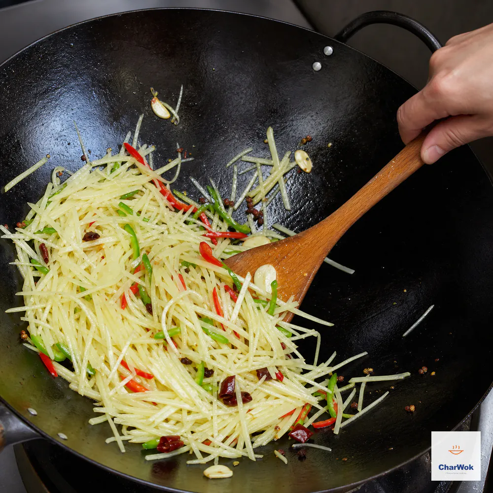 A wooden wok spatula tosses separate pale potato matchsticks with garlic, fresh red and green chili, dried chili, and peppercorns over high heat