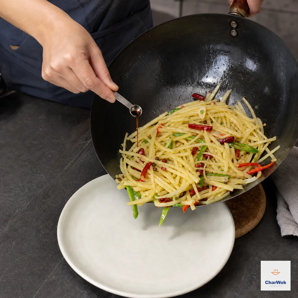 A tiny half-teaspoon measure drizzles the reserved Chinkiang vinegar over crisp potato strips in a wok resting off heat and tilted toward an empty plate