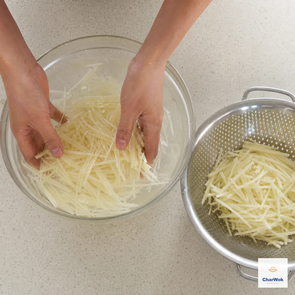 Two hands swish uniform 2-millimetre potato matchsticks in almost-clear cold water beside a colander of thoroughly drained strips