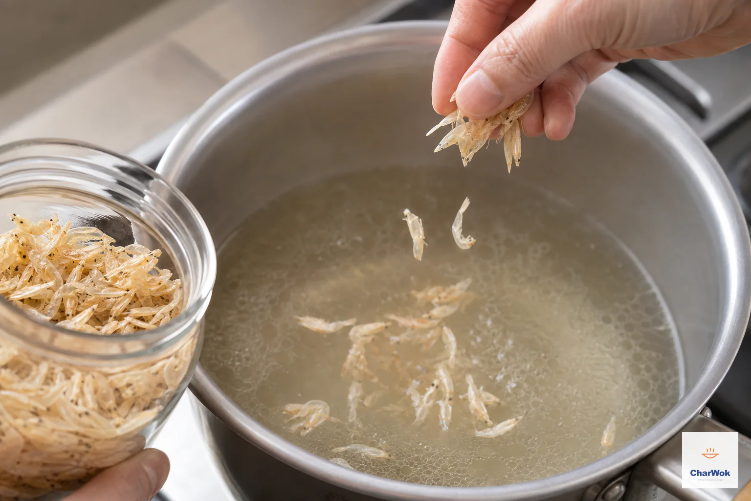Tiny pale translucent papery shrimp with intact heads, tails and shells are pinched straight from a dry jar into clear gently simmering broth