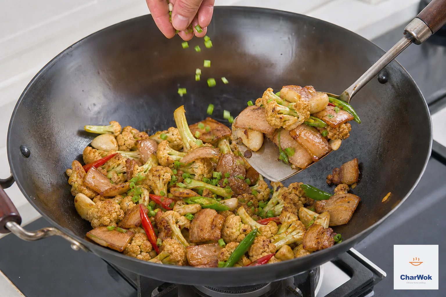 Chopped scallion falls over dry caramelized cauliflower and golden pork belly as a wok spatula lifts one final portion above an unlit burner