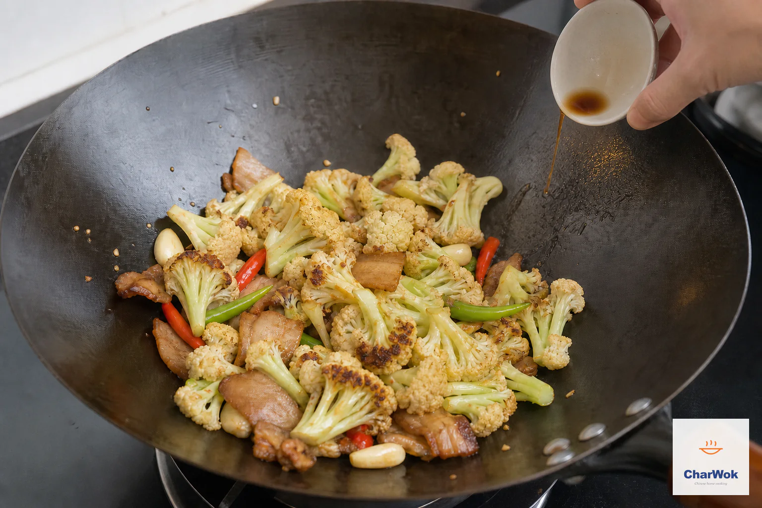 A one-teaspoon trickle of light soy runs down the exposed hot wok wall beside dry-tossed cauliflower with caramel patches, pork belly and chilies