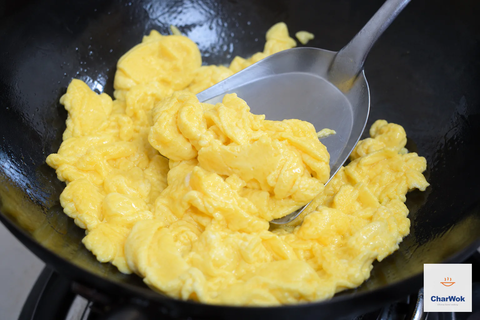 A metal spatula lifts tender pale-yellow scrambled egg curds from shimmering oil in a hot wok with no smoke present