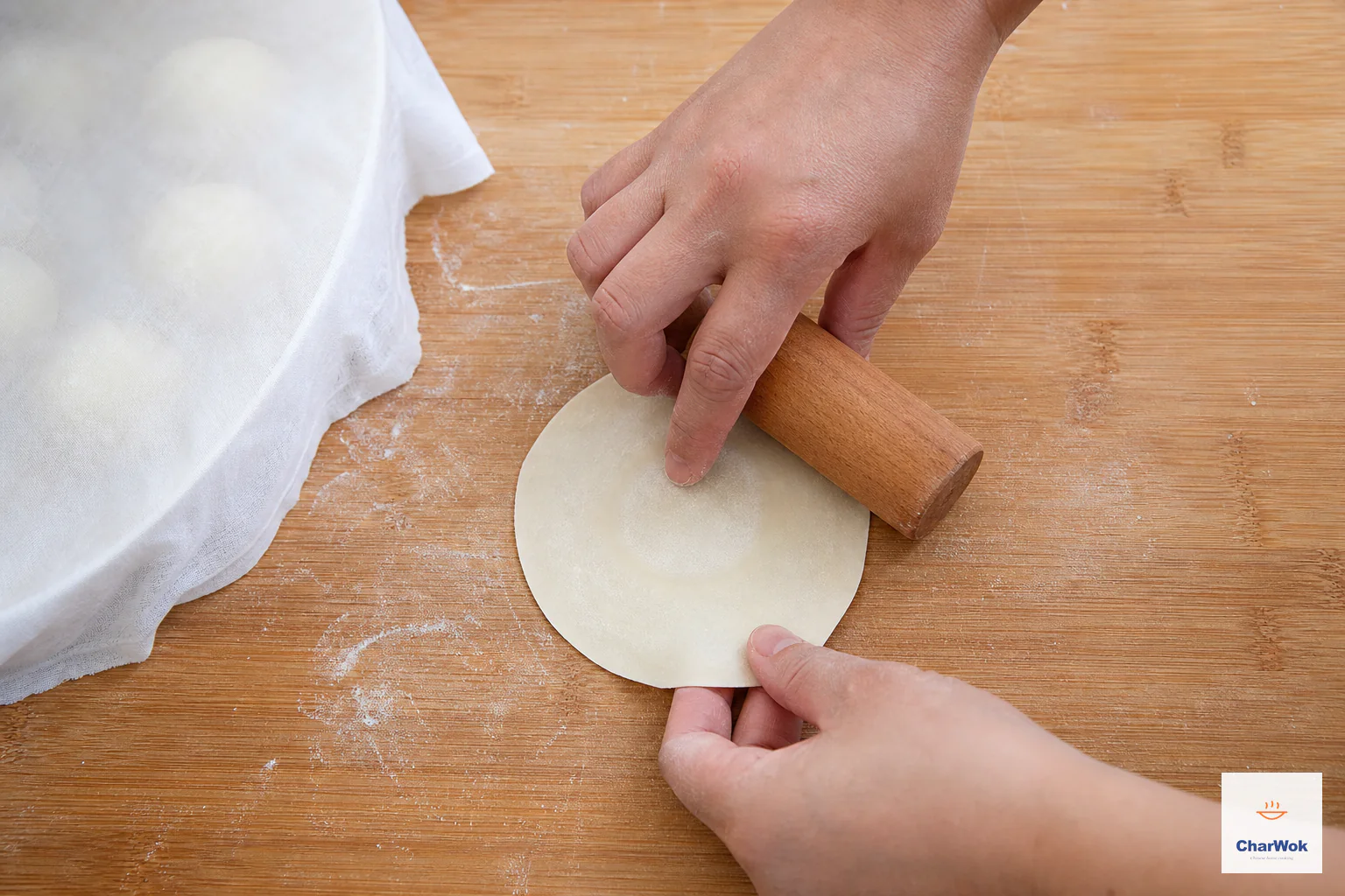 A short handleless rolling pin rolls the edge of a small round dumpling wrapper while one hand turns it, leaving the center visibly thicker