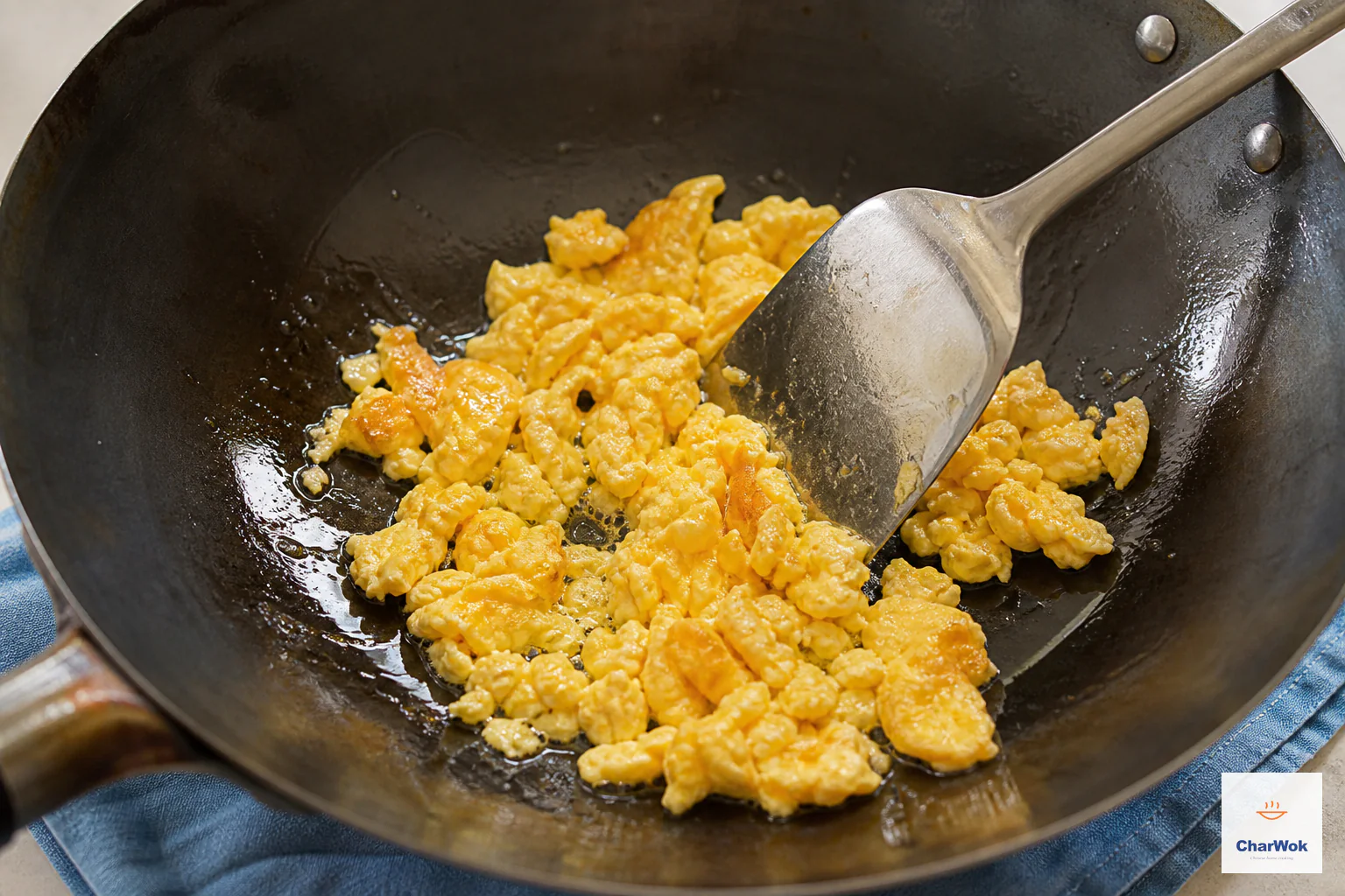 A metal wok spatula chops puffed egg into separate ragged golden curds with lightly browned edges