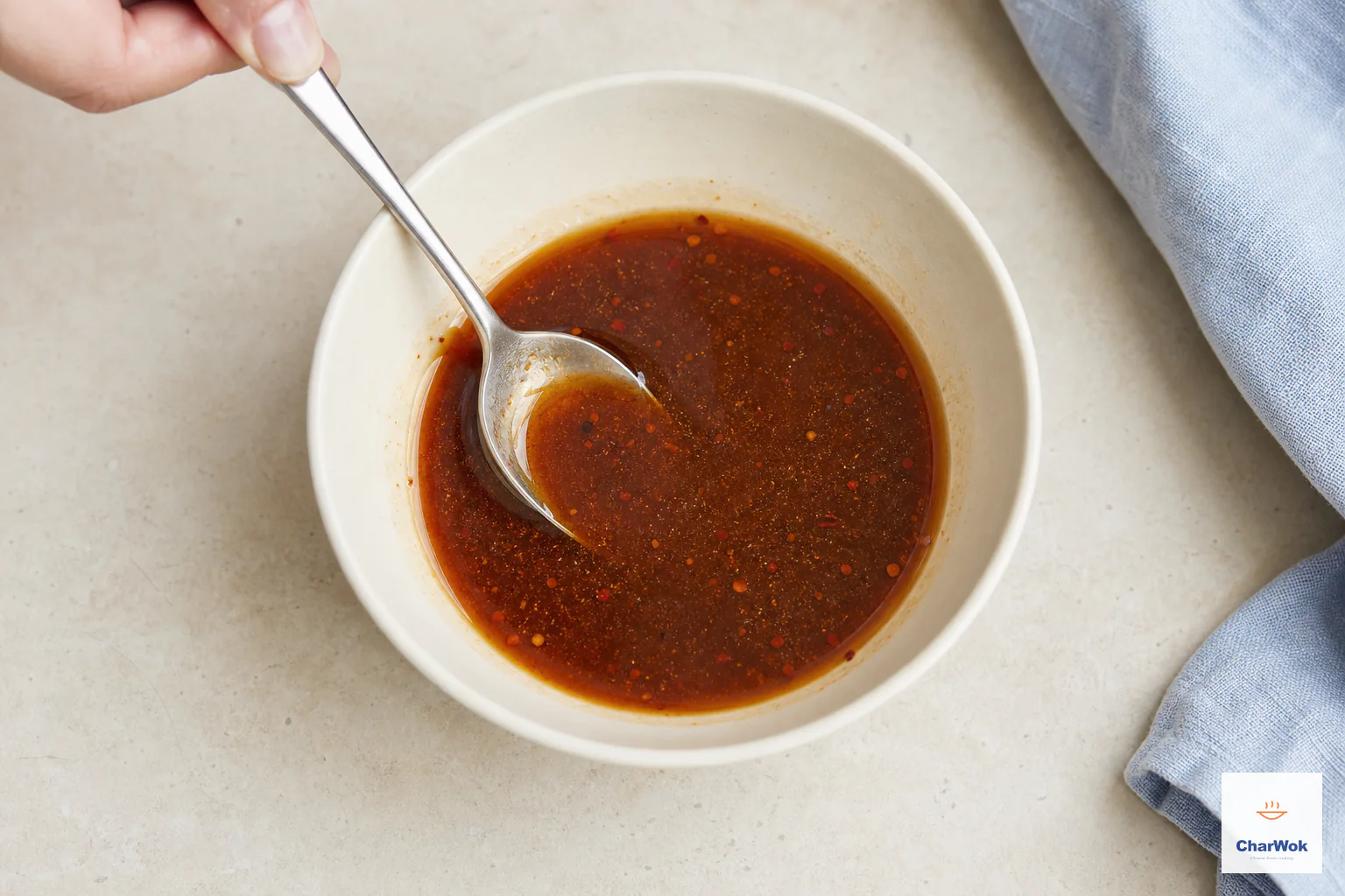 A spoon stirs a thin reddish-brown dressing with a light red-oil sheen and tiny chilli flakes in a cream bowl