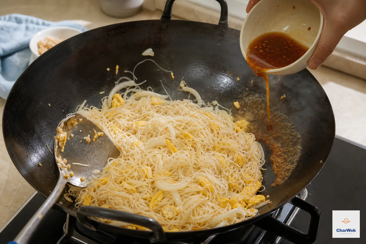 Reddish-brown dressing pours onto the exposed hot wok rim beside pale rice noodles, egg and onion