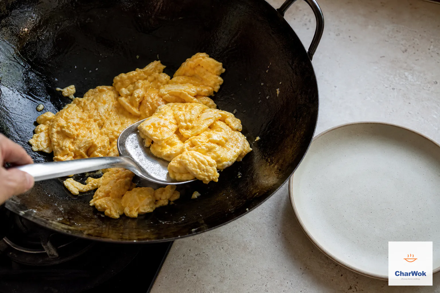 A metal spatula lifts large moist glossy golden egg curds from a dark wok toward a plain plate with no raw liquid pools remaining