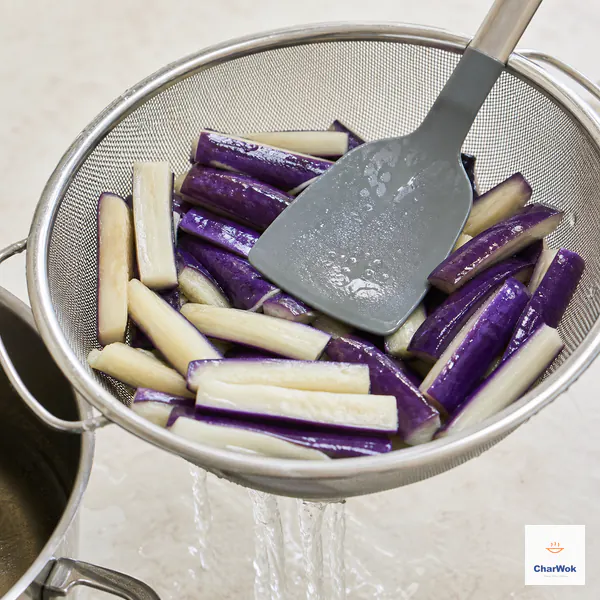 Vivid purple blanched eggplant batons drain in a metal colander while a broad spatula presses them gently
