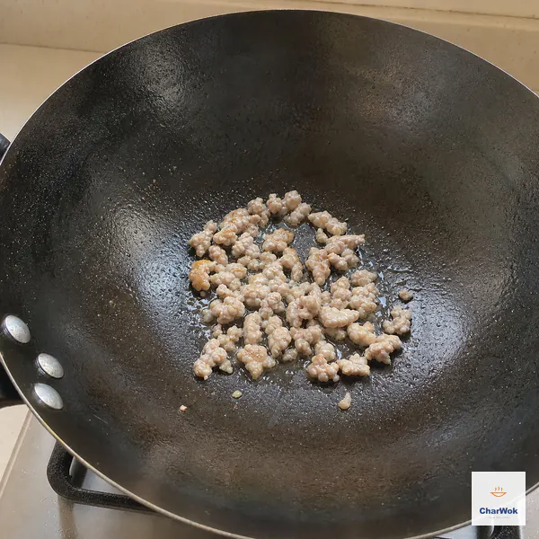 A modest portion of ground pork fries as small pale crumbles in a lightly oiled carbon-steel wok