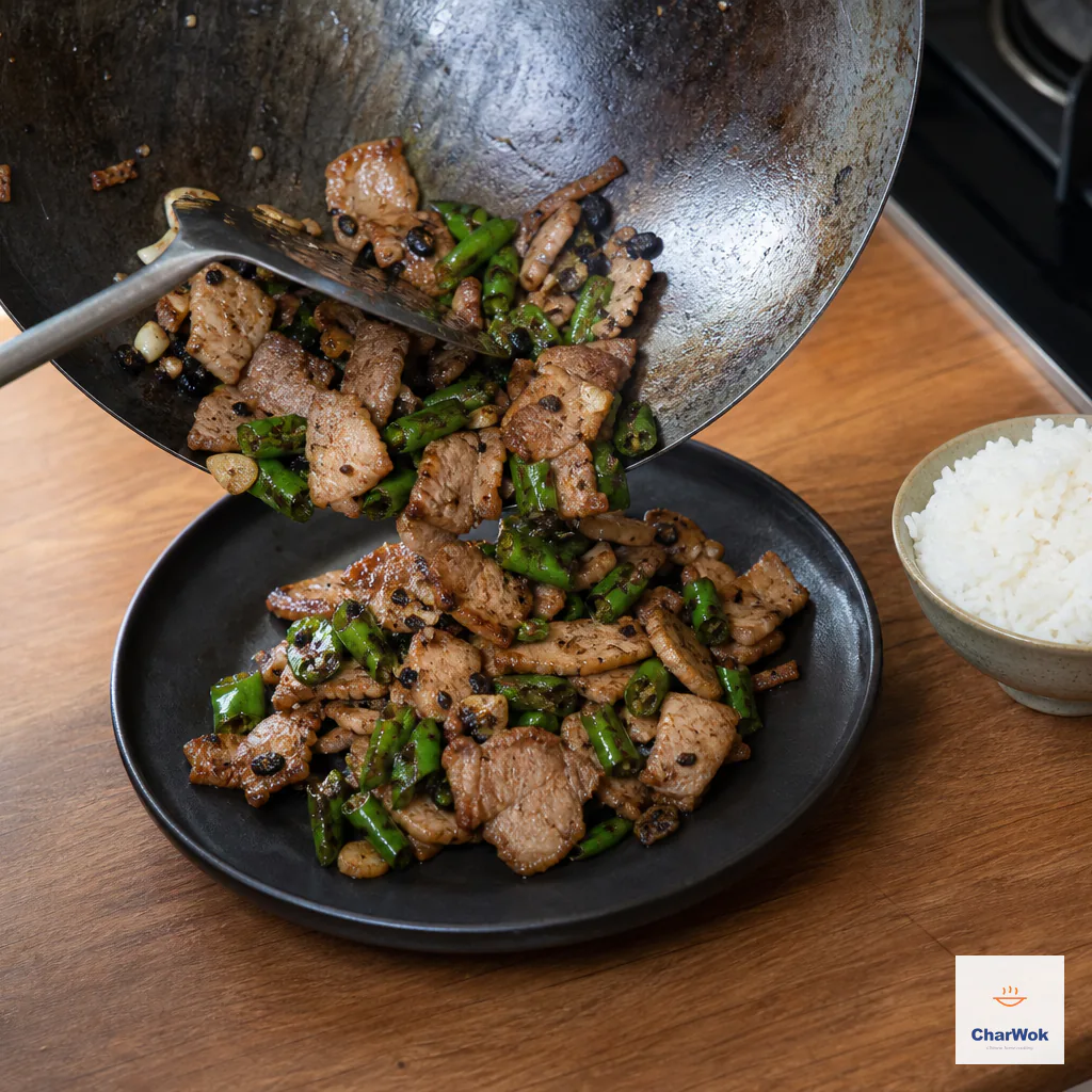 Dry Hunan pork stir-fry is scraped from the wok onto one dark plate beside a separate bowl of steamed jasmine rice
