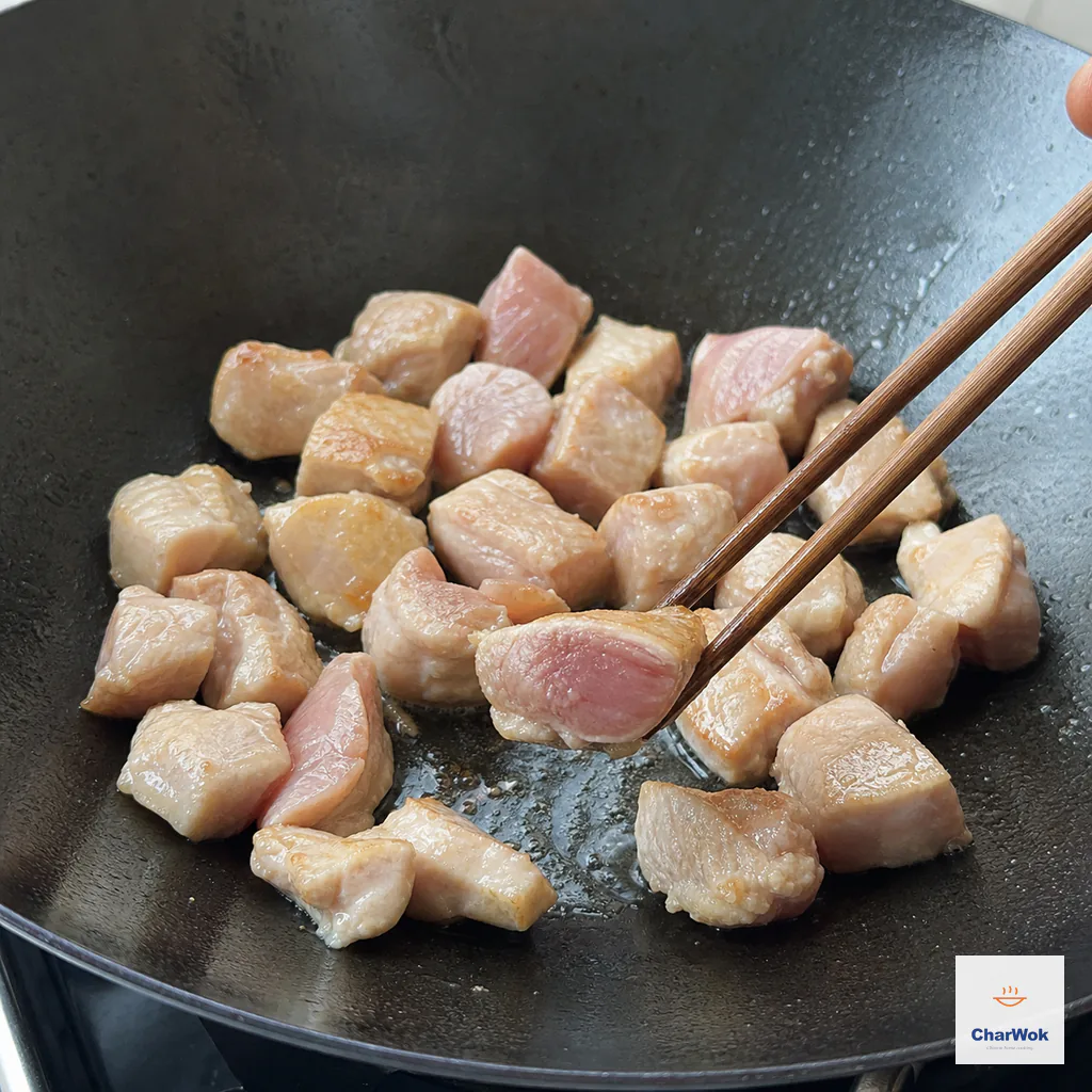 Chicken thigh pieces sear in a single layer in a wok while chopsticks hold one cut piece showing an opaque lightly golden outside and rosy raw center