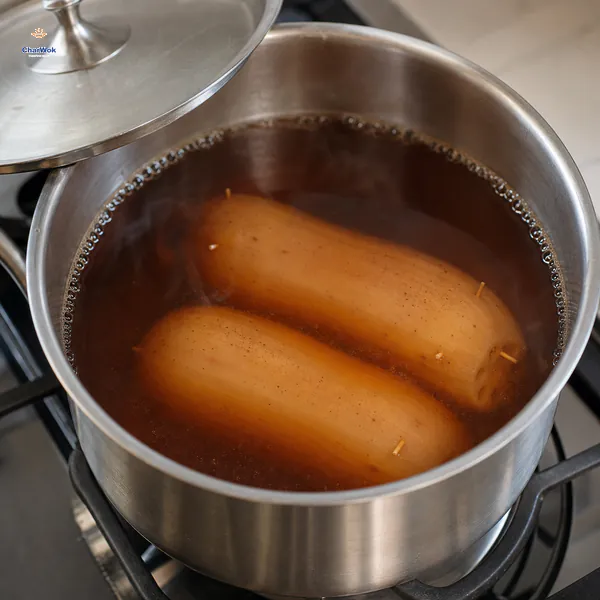 Two sealed lotus roots sit fully submerged in amber brown-sugar liquid at a very low simmer under a loosely set lid