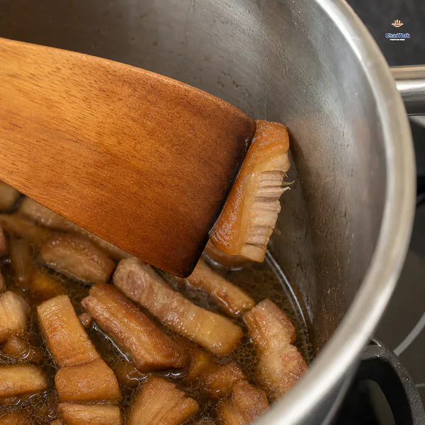 A wooden spatula gently presses one small braised pork belly strip against the pot wall as the softened skin and meat begin to part