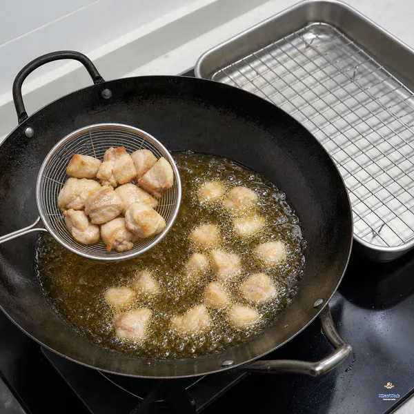 Uncoated chicken pieces fry in a wok while a wire skimmer lifts pieces that are still pale with only light-golden edges
